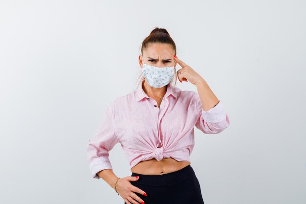 Young lady in shirt, mask keeping finger on temples and looking angry , front view.