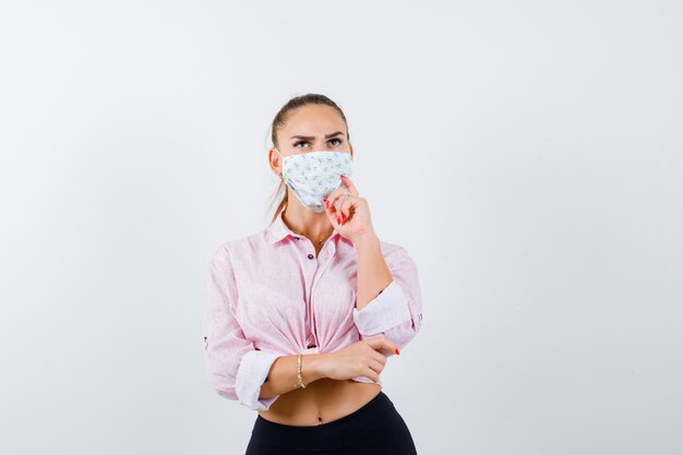 Young lady in shirt, mask keeping hand on chin and looking pensive , front view.