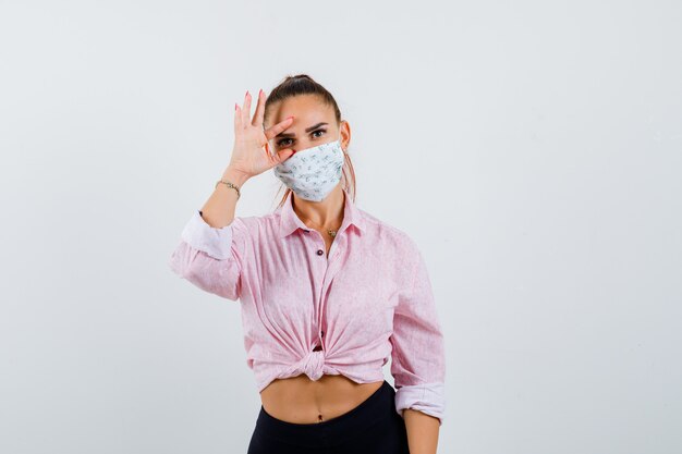 Young lady in shirt, mask opening eye with fingers and looking focused
