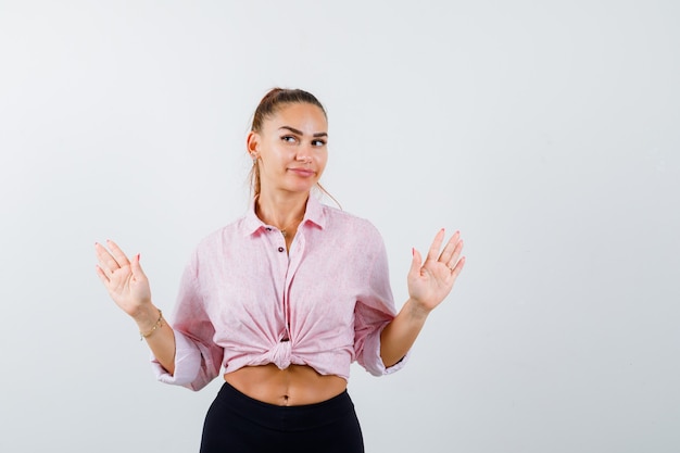 Young lady in shirt, pants showing palms in surrender gesture and looking focused , front view.