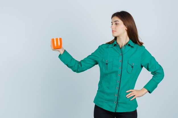 Young lady in shirt posing while holding orange cup of tea and looking confident , front view.