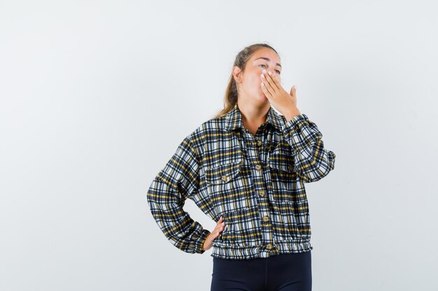 Giovane donna in camicia, pantaloncini tenendo la mano sulla bocca mentre sbadiglia e sembra assonnato, vista frontale.