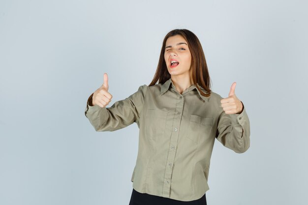 Молодая дама в рубашке, юбке показывает двойные пальцы вверх, моргая и выглядит счастливой, вид спереди.