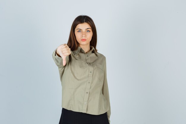 Young lady in shirt, skirt showing thumb down and looking disappointed , front view.