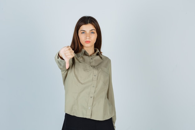 Молодая дама в рубашке, юбке показывает большой палец вниз и разочарованно смотрит, вид спереди.