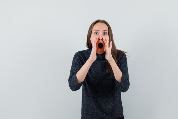 Young lady in shirt telling secret with hands near mouth and looking surprised