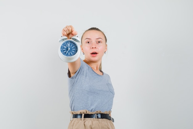 Young lady showing alarm clock in t-shirt and pants and looking jolly