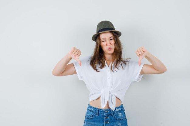 Young lady showing double thumbs down in t-shirt, jeans, hat and looking displeased , front view.