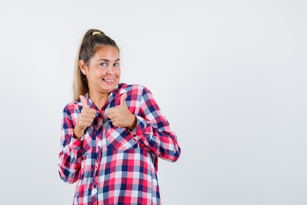 Young lady showing double thumbs up in checked shirt and looking happy. front view.