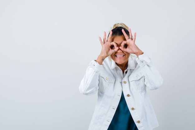 Young lady showing glasses gesture in shirt, white jacket and looking amused , front view.