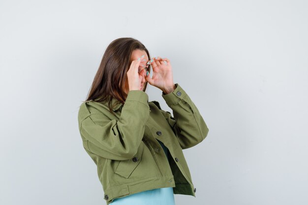 Young lady showing glasses gesture in t-shirt, jacket and looking funny. front view.