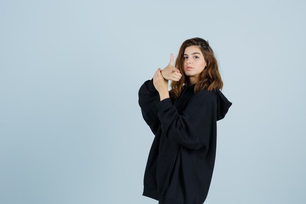 Young lady showing gun gesture in oversized hoodie, pants and looking serious , front view.