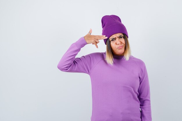 Young lady showing gun gesture in purple sweater, beanie and looking bored. front view.
