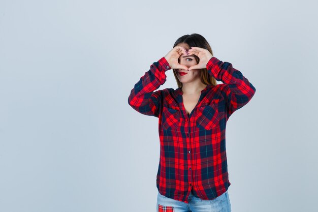 Young lady showing heart gesture in checked shirt and looking confident. front view.