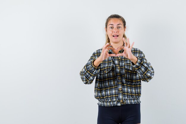 Young lady showing heart gesture in checked shirt, shorts and looking merry. front view.