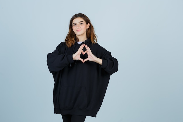 Young lady showing heart gesture in oversized hoodie, pants and looking cute. front view.