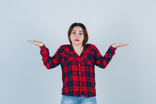 Young lady showing helpless gesture in checked shirt, jeans and looking indecisive , front view.