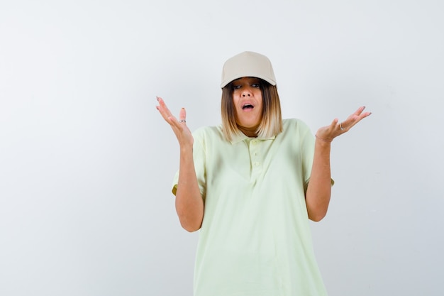 Young lady showing helpless gesture in t-shirt, cap and looking stressed. front view.