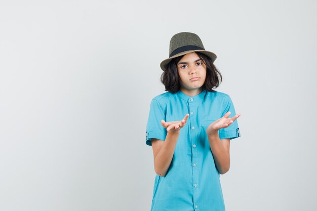 파란색 셔츠, 모자에 손으로 무기력 한 제스처를 보여주는 젊은 아가씨 감정적 찾고