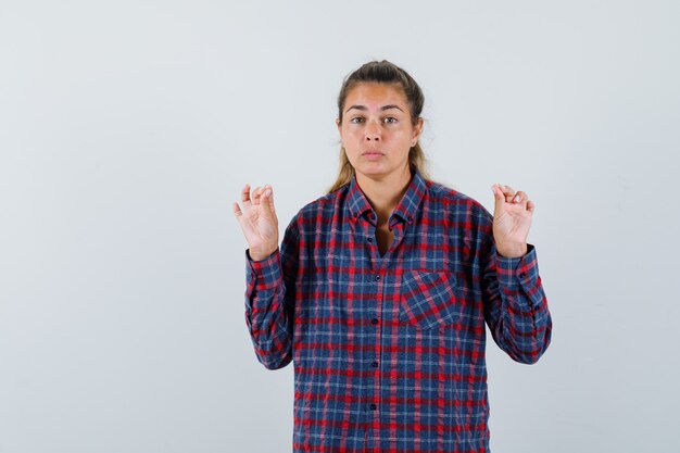 Young lady showing meditation gesture in checked shirt and looking hopeful. front view.