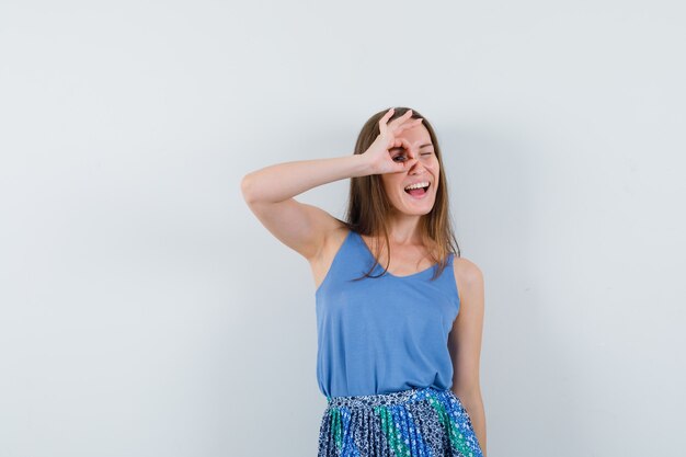 Young lady showing ok gesture on eye in singlet, skirt and looking merry. front view.