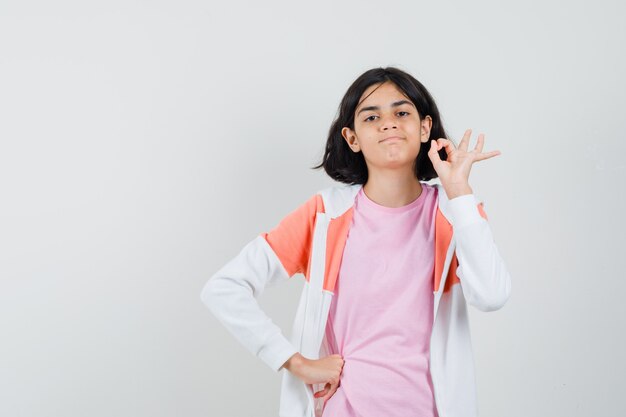 Young lady showing ok gesture in jacket, pink shirt and looking satisfied