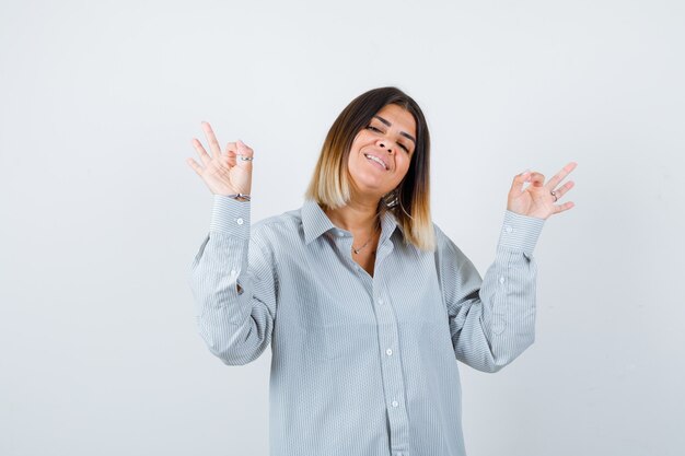 Young lady showing ok gesture in oversize shirt and looking joyful. front view.