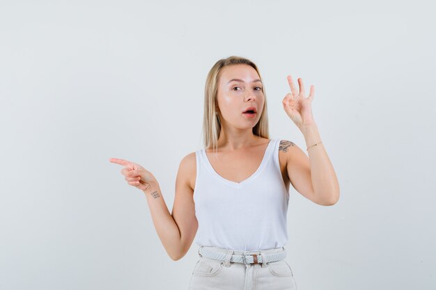 Young lady showing ok gesture while pointing aside in white blouse and looking assured