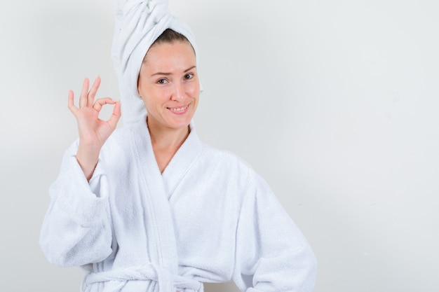 Young lady showing ok gesture in white bathrobe, towel and looking happy. front view.