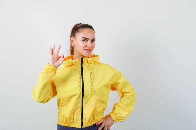 Young lady showing ok gesture in yellow jacket and looking pleased , front view.