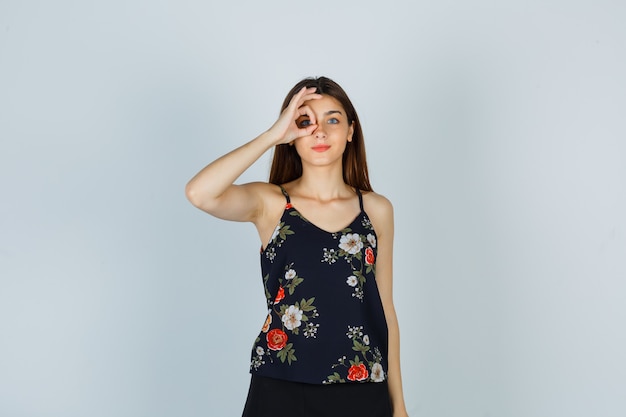 Young lady showing ok sign on eye in blouse and looking confident. front view.