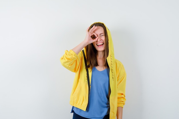 Young lady showing ok sign on eye in t-shirt, jacket and looking glad, front view.