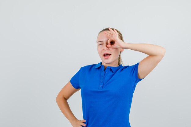 Young lady showing ok sign on eye while winking in blue t-shirt