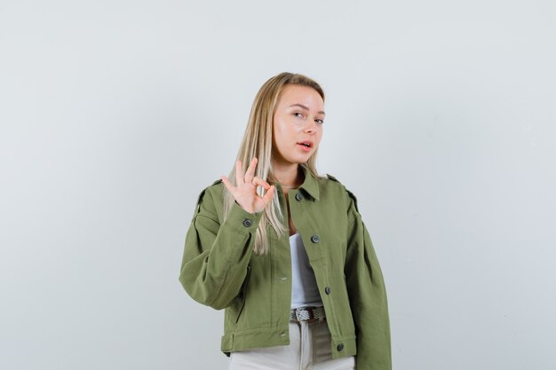 ジャケット、パンツでOKサインを示し、自信を持って見える若い女性。正面図。