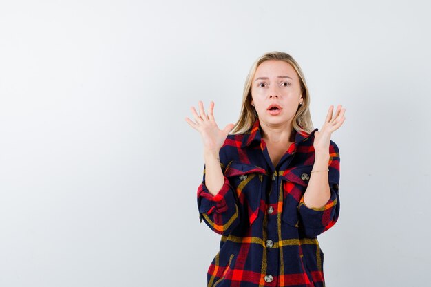 Young lady showing palms in surrender gesture in checked shirt and looking scared , front view.