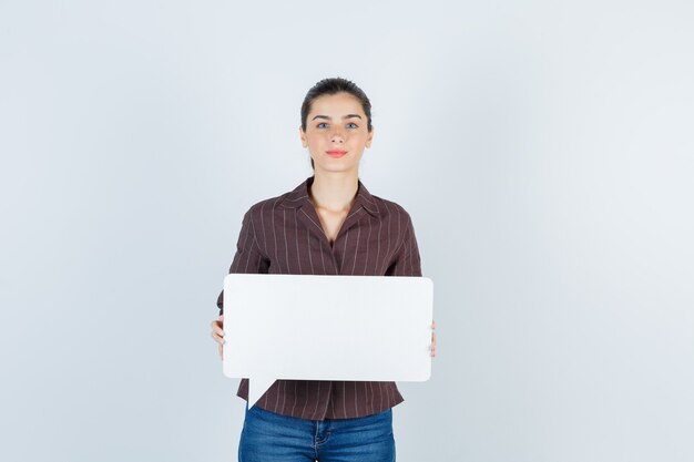 Young lady showing paper poster in shirt, jeans and looking serious , front view.