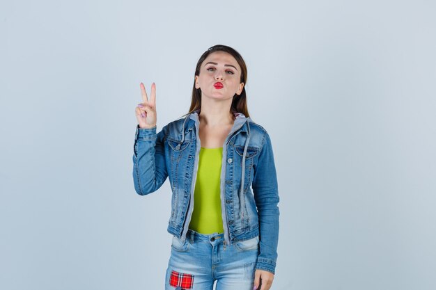 Young lady showing peace gesture, pouting lips in denim outfit and looking cute. front view.