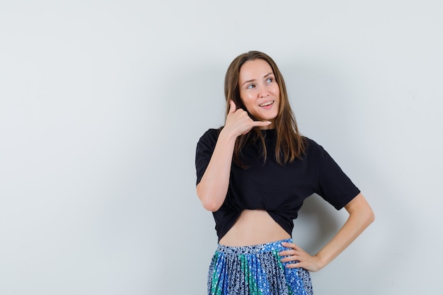 Young lady showing phone gesture in blouse, skirt and looking dreamy