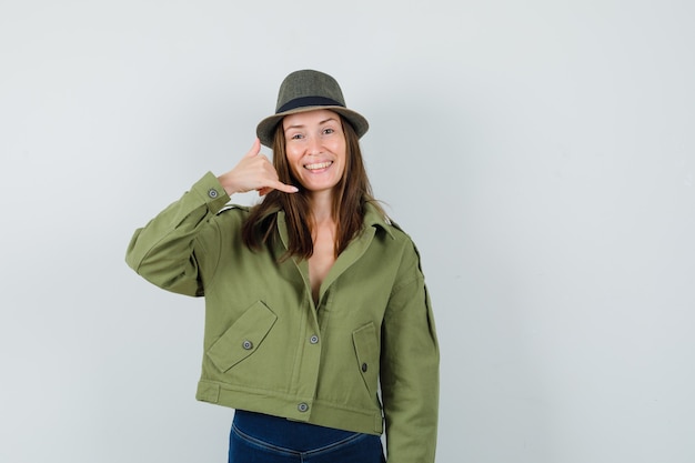 Young lady showing phone gesture in jacket pants hat and looking cheerful  