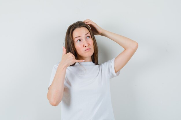 Young lady showing phone gesture while scratching head in white t-shirt and looking forgetful 