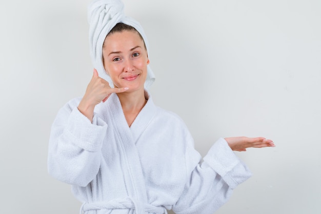 Young lady showing phone gesture in white bathrobe, towel and looking pleased , front view.