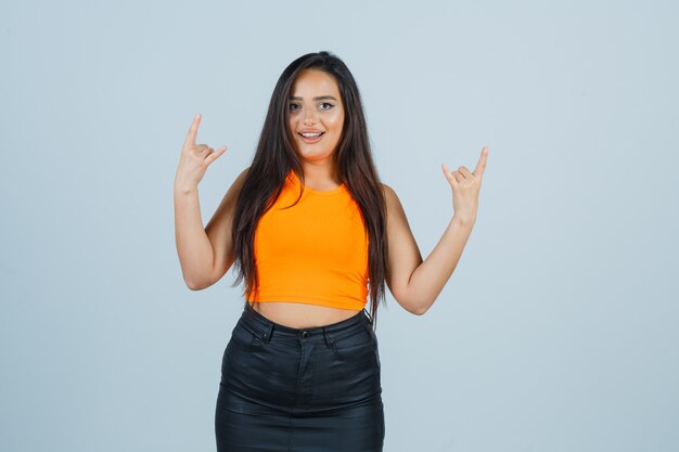 Young lady showing rock gesture in singlet, mini skirt and looking crazy. front view.