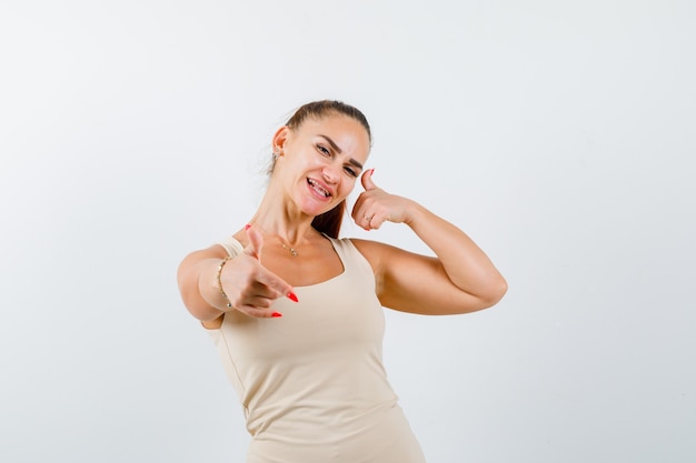 Young lady showing rock gesture and thumb up in tank top and looking glad , front view.