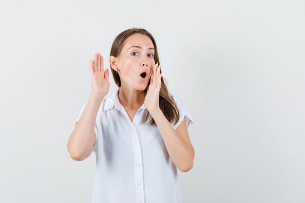 Young lady showing shouting while listening gesture in white blouse and looking focused