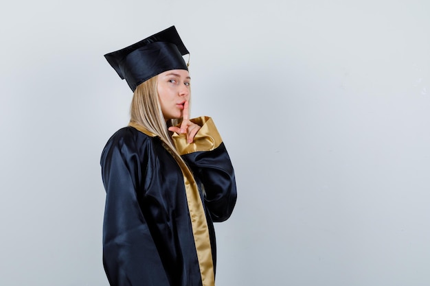 Young lady showing silence gesture in academic dress and looking confident.