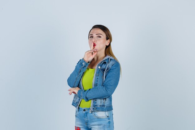 Young lady showing silence gesture in denim outfit and looking mysterious. front view.