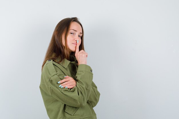 Young lady showing silence gesture in green jacket and looking mysterious , front view.