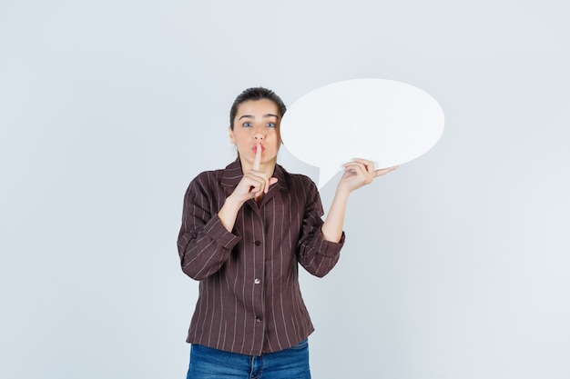 Young lady showing silence gesture, keeping paper poster in shirt, jeans and looking serious , front view.