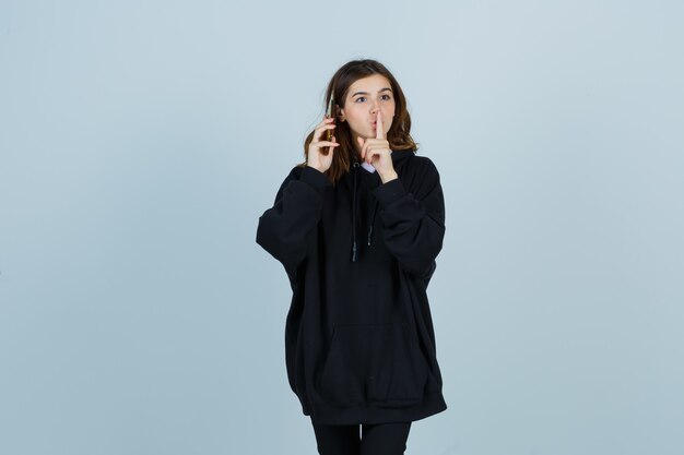 Young lady showing silence gesture in oversized hoodie, pants and looking serious. front view.