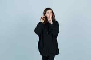 Free photo young lady showing silence gesture in oversized hoodie, pants and looking serious. front view.
